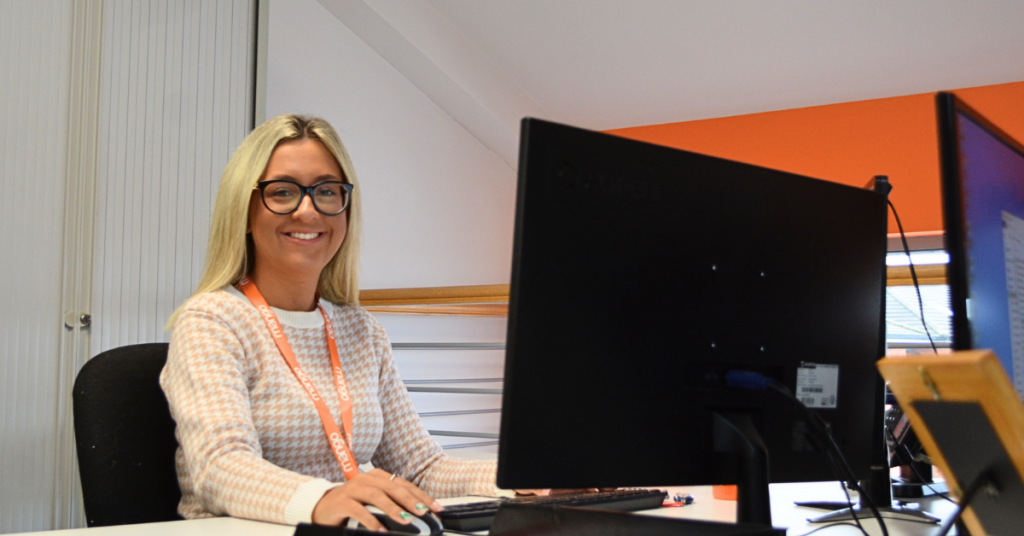 Mango Direct Marketing employee sitting at desk in front of computer smiling, blonde hair, orange lanyard, live chat service